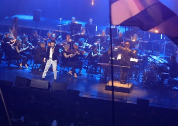 mężczyzna śpiewający na scenie za nim orkiestra symfoniczna